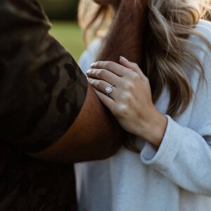 May include: A hand wearing a silver engagement ring with a round center stone and a halo of smaller stones. The ring is on a person's finger, with another person's arm in the foreground. The background is blurred.