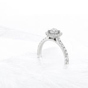 Pode incluir: Um anel de noivado prateado com uma pedra central de corte quadrado rodeada por um halo de pedras menores. A banda tamb&eacute;m &eacute; adornada com pequenas pedras brilhantes. O anel est&aacute; sobre um fundo branco suave.