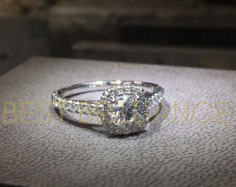 Op de afbeelding: Een zilveren ring met een grote, vierkant geslepen middensteen omringd door een halo van kleinere stenen. De band is ook versierd met kleinere stenen. De ring ligt op een grijze ondergrond.