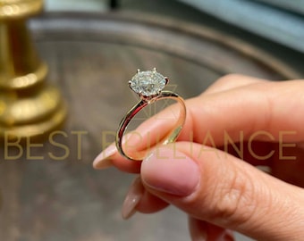 Pode incluir: Um anel de noivado de ouro com um grande diamante transparente de corte redondo. O anel é segurado por uma mão. A banda do anel é fina e polida. O texto "BEST BRILLIANCE" é visível no fundo.