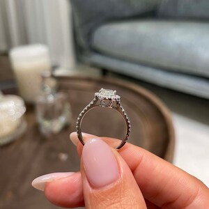 May include: A silver engagement ring with a square-cut center stone and a halo of smaller stones. The band is also adorned with small stones. The ring is held up, showcasing its design and sparkle.