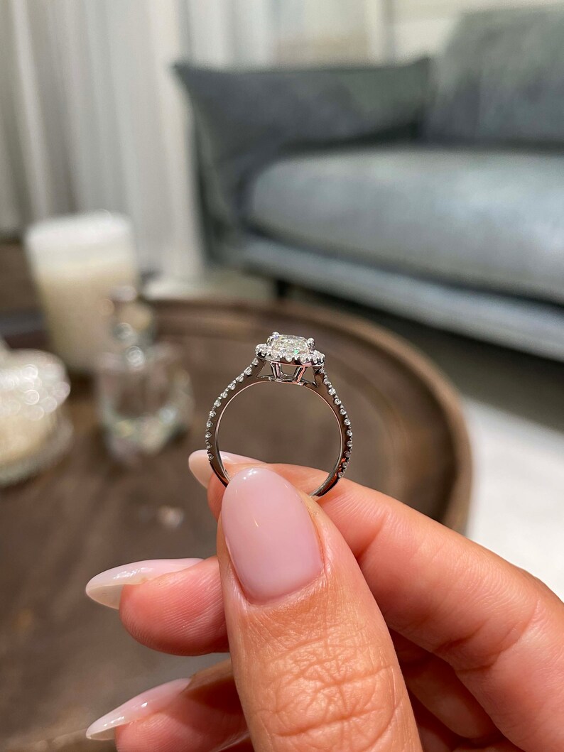May include: A silver engagement ring with a square-cut center stone and a halo of smaller stones. The band is also adorned with small stones. The ring is held up, showcasing its design and sparkle.