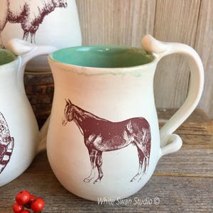 May include: White ceramic mug with a brown horse illustration and a light green interior. The mug has a unique handle with a loop at the top.