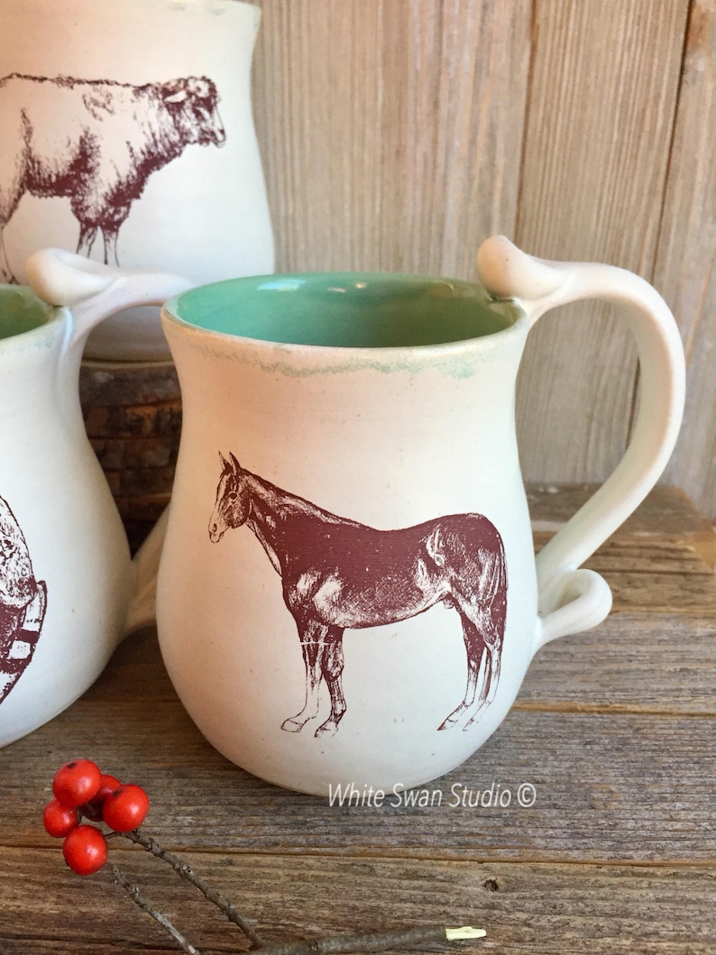 May include: White ceramic mug with a brown horse illustration and a light green interior. The mug has a unique handle with a loop at the top.