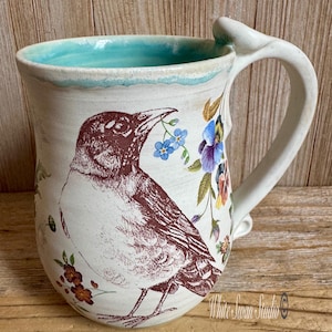 May include: Cream-colored ceramic mug with a bird illustration and floral accents. The bird is brown, holding a blue flower in its beak. The mug has a turquoise interior and a unique handle. The mug is from White Swan Studio.