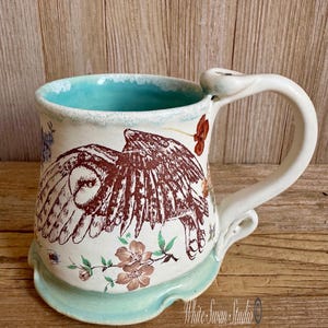 May include: A handmade ceramic mug with a white base, a turquoise interior, and a matching turquoise base. The mug features a brown owl design with floral accents. The handle is white and curved. The mug is decorated with floral accents.