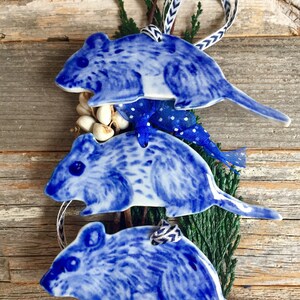 May include: Three blue and white ceramic mouse ornaments hanging from a branch. The mice are stacked on top of each other, with the largest mouse at the bottom and the smallest mouse at the top. The ornaments are decorated with a blue and white pattern.