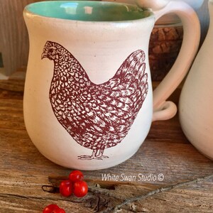 May include: White ceramic mug with a brown illustration of a hen on a white background. The mug has a light green interior and a white handle. The mug is sitting on a wooden surface with red berries.