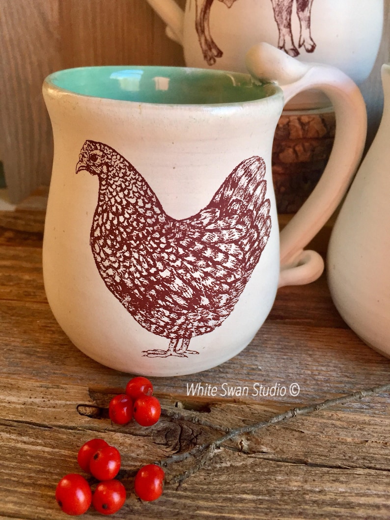 May include: White ceramic mug with a brown illustration of a hen on a white background. The mug has a light green interior and a white handle. The mug is sitting on a wooden surface with red berries.