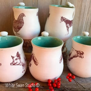 May include: Set of five ceramic mugs with a rustic farmhouse theme. Each mug features a different animal illustration: a chicken, a horse, two pigs, and a cow. The mugs have a white glaze with a light pink hue and a turquoise blue interior.