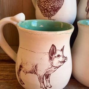 May include: A white ceramic mug with a teal green interior and a brown line around the rim. The mug features a brown line drawing of a pig on the front. The text "White Swan Studio &copy;" is printed on the bottom of the mug.