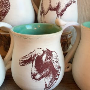 May include: White ceramic mug with a brown goat illustration and a turquoise blue glaze inside. The mug has a rounded body and a handle.