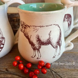 May include: White ceramic mug with a teal glaze interior and a brown illustration of a sheep. The mug has a unique bone-shaped handle. The mug is sitting on a wooden surface with red berries.