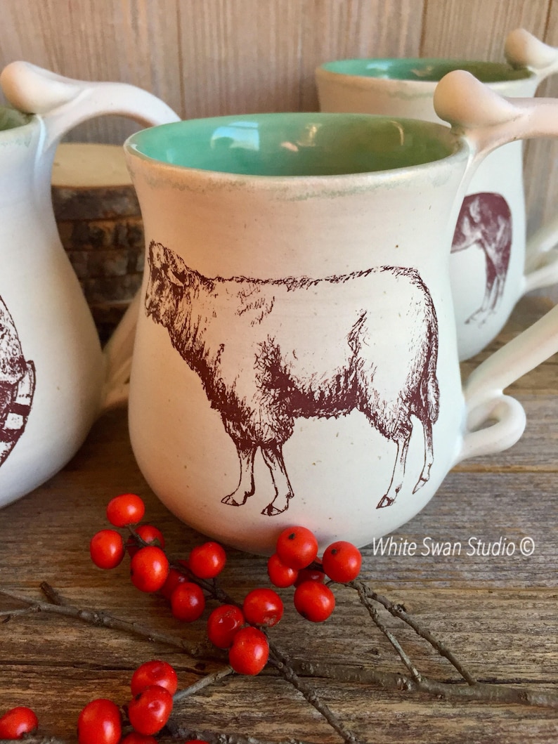May include: White ceramic mug with a teal glaze interior and a brown illustration of a sheep. The mug has a unique bone-shaped handle. The mug is sitting on a wooden surface with red berries.