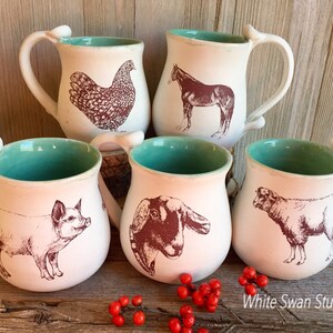 May include: Set of five ceramic mugs with a turquoise glaze and white interior. Each mug features a different farm animal illustration in brown ink: a chicken, a horse, a pig, a goat, and a sheep. The mugs are handmade and have a unique, rustic look.
