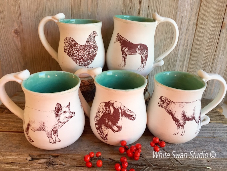 May include: Set of five ceramic mugs with a turquoise glaze and white interior. Each mug features a different farm animal illustration in brown ink: a chicken, a horse, a pig, a goat, and a sheep. The mugs are handmade and have a unique, rustic look.