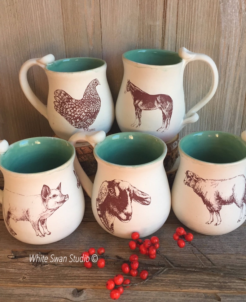 May include: Set of four white ceramic mugs with a turquoise rim. Each mug features a different farm animal illustration in brown ink: a chicken, a horse, a pig, and a goat. The mugs are arranged on a wooden surface with red berries.