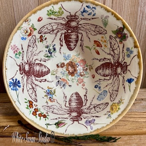 May include: A cream-coloured ceramic bowl with four large, detailed reddish-brown bee illustrations. The bowl is decorated with colourful floral patterns and has a distressed, gold-toned rim. The bowl's diameter is approximately 25 cm.