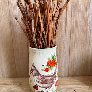 May include: A white ceramic vase with a bird and floral design, holding a bundle of brown, dried stems. The vase features a detailed illustration of a bird, red roses, and other flowers. The vase is on a wooden surface.