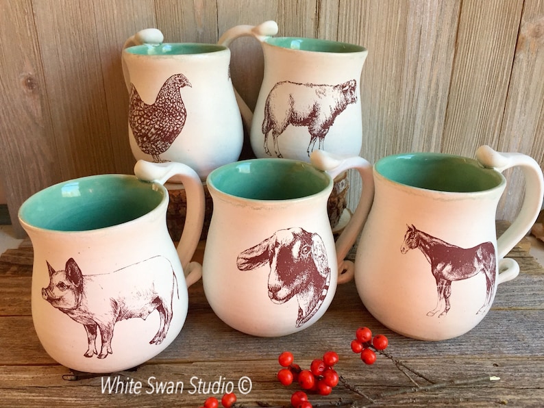 May include: Five ceramic mugs with a teal glaze and white interior. Each mug features a different farm animal illustration in brown ink: a pig, a goat, a horse, a sheep, and a chicken. The mugs have a rounded shape and a handle.