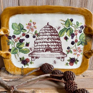 May include: Rectangular serving tray with a cream-colored center featuring a beehive illustration, surrounded by floral and berry motifs. The tray has a gold-colored border and handles, and is placed on a wooden surface.