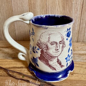 May include: A ceramic mug featuring a brown portrait of George Washington on a cream-colored background, accented with blue floral patterns. The mug has a deep blue interior, a scalloped blue base, and a uniquely shaped, curved white handle.