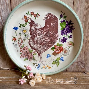 May include: A round, off-white decorative plate with a light teal rim. The plate features a detailed illustration of a brown hen surrounded by colorful flowers. The text "White Swan Studio" is visible on the plate.