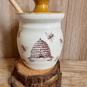 May include: A cream-colored ceramic honey pot with a brown beehive and bee illustrations. The pot has a yellow lid and includes a wooden honey dipper. The pot is displayed on a wooden slice.