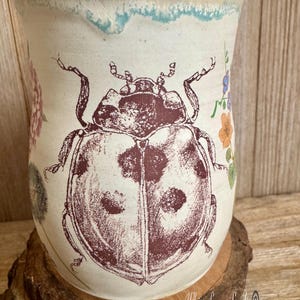 May include: Cream-colored ceramic mug featuring a large maroon ladybug illustration. The mug has a slightly textured surface with floral and blue accents near the rim. The mug is sitting on a wooden base.