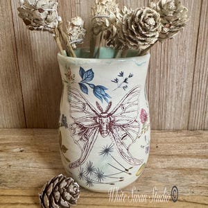 May include: A ceramic vase with a moth and floral design, holding dried pine cones and stems. The vase is off-white with a light blue interior and a brown moth illustration. A single pine cone rests in front of the vase.
