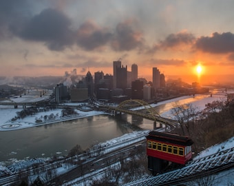 Puede incluir: Un funicular rojo de estilo vintage asciende por una colina cubierta de nieve con vistas al horizonte de una ciudad al amanecer. La ciudad está cubierta de nieve y el río está congelado. El sol sale por detrás de la ciudad, proyectando un cálido resplandor sobre la escena.