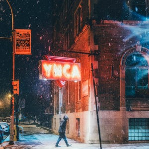 May include: A red neon sign with the letters YMCA on a brick building in a snowy city at night. The sign is illuminated and there is a person walking in the street.