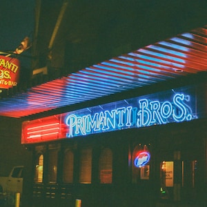 May include: A neon sign for Primanti Bros. Restaurant & Bar with blue neon letters on a clear background. The sign is lit up and there are red neon lights above it. The sign is mounted on a building with a striped awning.