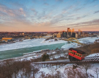 Puede incluir: Un funicular rojo y amarillo asciende por una colina nevada con vistas a un río helado y el horizonte de una ciudad. La ciudad está cubierta de nieve y el cielo es de un suave rosa y azul.