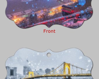 May include: Ornament with a winter cityscape on the front, featuring a city skyline at night with lights and a funicular. The back shows a bridge over water in a black and white winter scene.