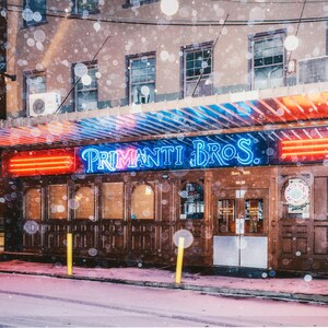 May include: A storefront with a neon sign that reads "Primanti Bros." The sign is lit up in pink and blue. The storefront is covered in snow.