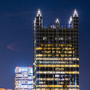 May include: A tall, modern skyscraper with a distinctive pointed roofline, illuminated at night. The building is made of glass and steel, with a grid pattern of windows. The building is lit up with warm yellow lights, creating a striking contrast against the dark blue sky. The building is located in a city, and other buildings can be seen in the background.