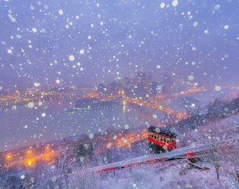 May include: A red cable car ascends a snowy hill with a view of a city skyline in the distance. Snow falls softly over the city and the river below.