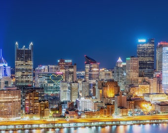 Puede incluir: Una vista nocturna del horizonte de Pittsburgh con edificios altos iluminados en varios colores. Los edificios están agrupados y se reflejan en el agua de abajo.