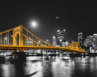 Puede incluir: Un puente amarillo cruza un río frente a un horizonte urbano nocturno. El puente está iluminado y el horizonte urbano está en blanco y negro.
