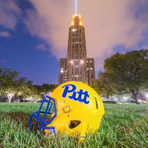 May include: A yellow football helmet with blue accents and the word "Pitt" in blue lettering rests on a grassy field. In the background, a tall stone building is illuminated at night, with a beam of light emanating from the top.
