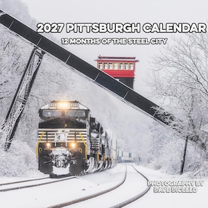 May include: A 2027 Pittsburgh calendar titled 12 Months of the Steel City featuring a photograph of a black and white freight train moving along snowy tracks beneath a red structure and snow-covered trees, with photography by Dave Dicello.