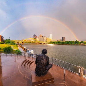 May include: A bronze sculpture of a person sitting on a platform overlooking a river with a double rainbow in the background. The city skyline is visible in the distance.