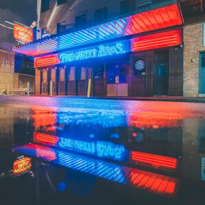 May include: A neon sign for Primanti Bros. restaurant with blue and red lights. The sign is reflected in a puddle of water on the street.