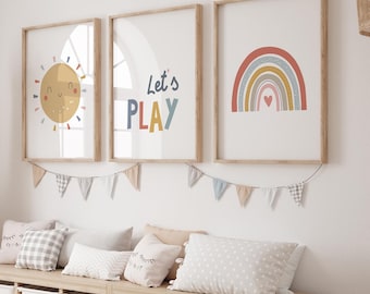May include: Three framed art prints with a sun, rainbow, and the words "Let's PLAY" in a child's room. The prints are in wooden frames and are hanging above a bench with pillows and a garland of pennants.
