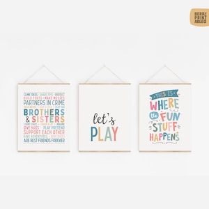 Puede incluir: Tres impresiones artísticas enmarcadas con colgadores de madera. Las impresiones presentan texto colorido: "Brothers & Sisters", "let's PLAY" y "This is where the FUN stuff HAPPENS". Las impresiones están sobre un fondo blanco.