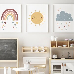 Könnte beinhalten: Drei druckbare Wandbilder mit einem Regenbogen mit Herzen, einer lächelnden Sonne und einer blauen Wolke mit Sternen. Die Drucke sind an einer weißen Wand in einem Kinderspielzimmer angebracht.
