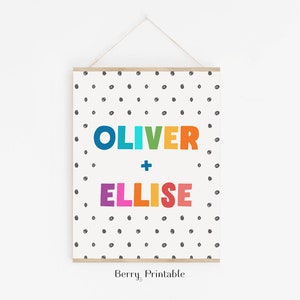 Op de afbeelding: Een ingelijste print met de namen "OLIVER" en "ELLISE" in kleurrijke letters. De namen staan centraal op een witte achtergrond met verspreide zwarte stippen. De print wordt opgehangen met een houten balk en touw.