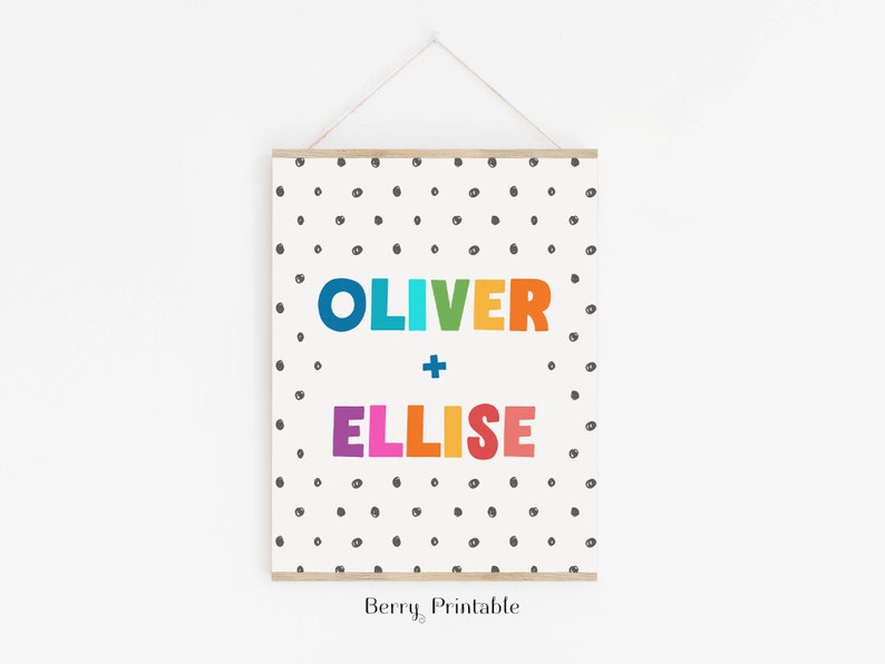 Op de afbeelding: Een ingelijste print met de namen "OLIVER" en "ELLISE" in kleurrijke letters. De namen staan centraal op een witte achtergrond met verspreide zwarte stippen. De print wordt opgehangen met een houten balk en touw.