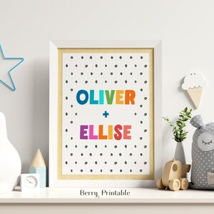 Op de afbeelding: Een ingelijste print met de namen "OLIVER + ELLISE" in kleurrijke letters op een witte achtergrond met zwarte stippen. De print staat op een witte plank met een ster, een konijn en een beer, ideaal voor een kinderkamer.